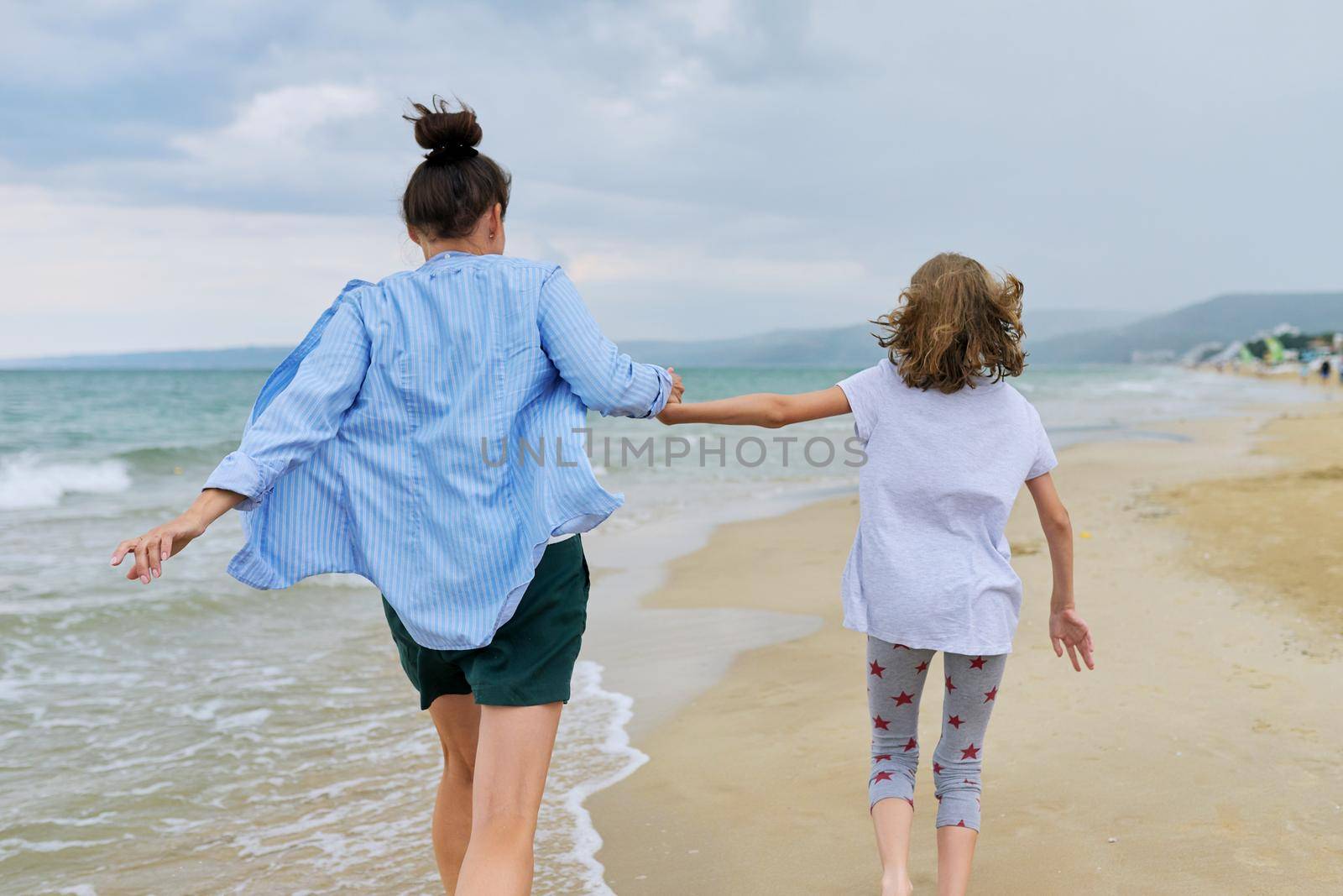Happy mom and daughter running on the sea beach holding hands, back view by VH-studio