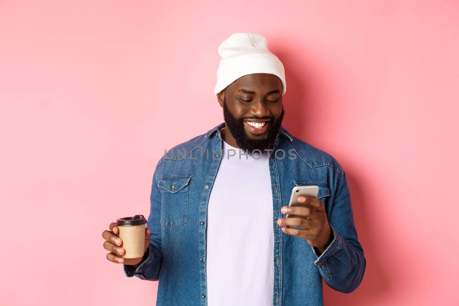 Image of stylish Black man hipster drinking takeaway coffee, reading message on phone and smiling, standing over pink background by Benzoix