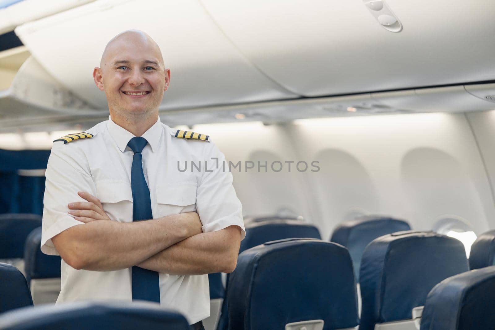 Cheerful male pilot in uniform keeping arms crossed and smiling at camera while standing inside of the airplane. Transportation, aircrew concept