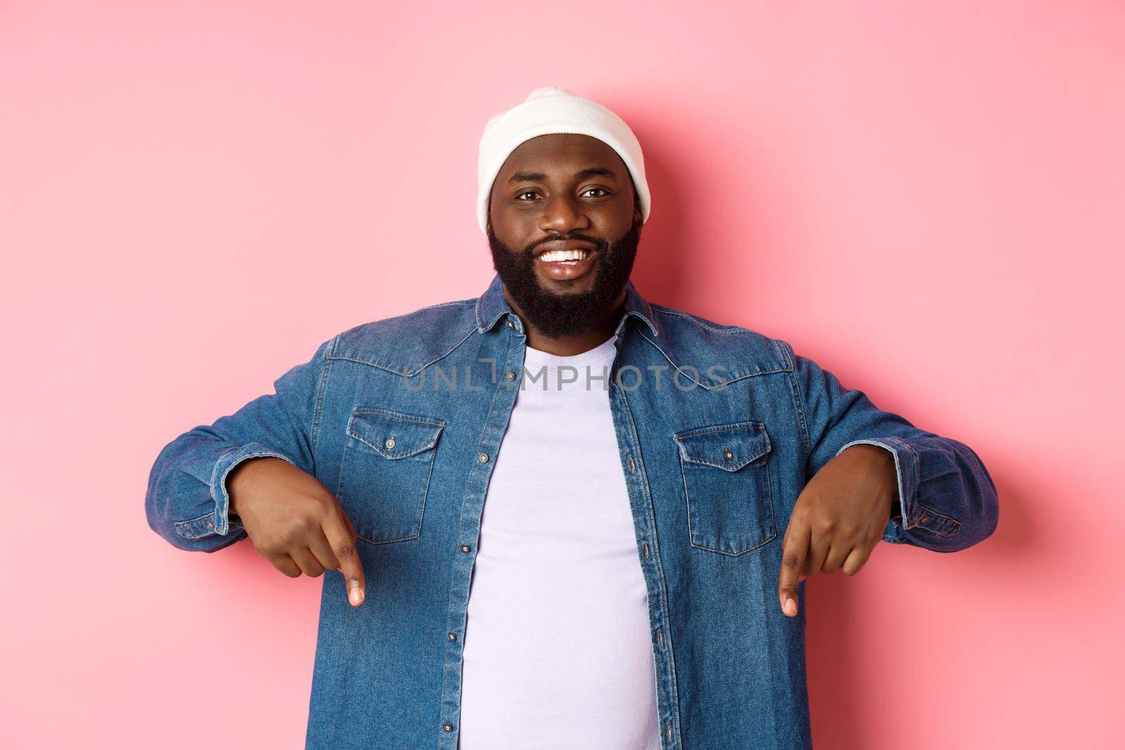 Handsome hipster african-american man pointing fingers down, smiling and showing promo offer, standing over pink background.