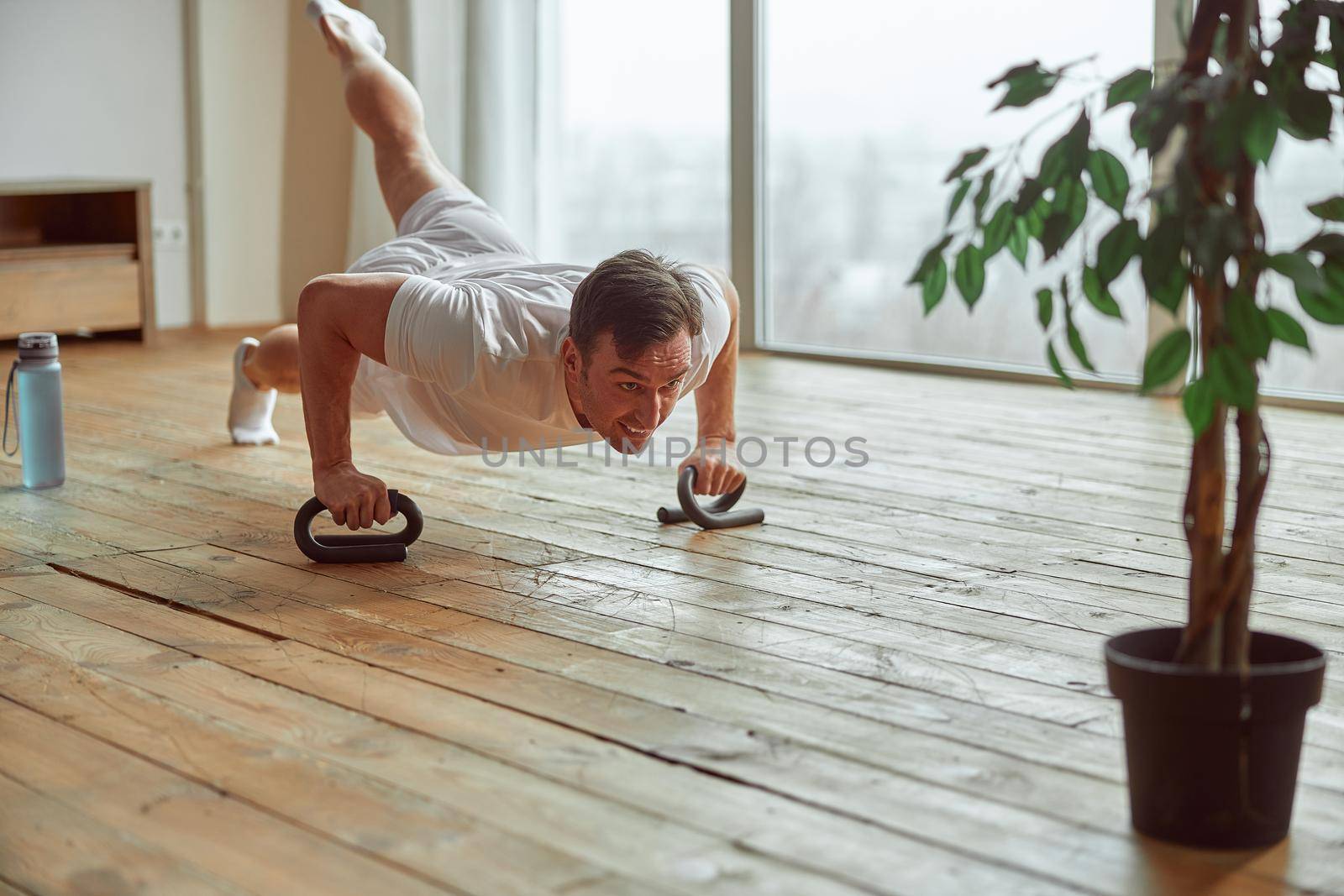 Motivated male is doing push up on one foot while using special handles for balancing near window