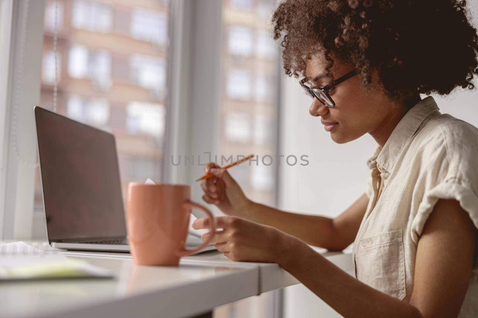 Pretty Afro American woman working with laptop and looking on document while sitting near window