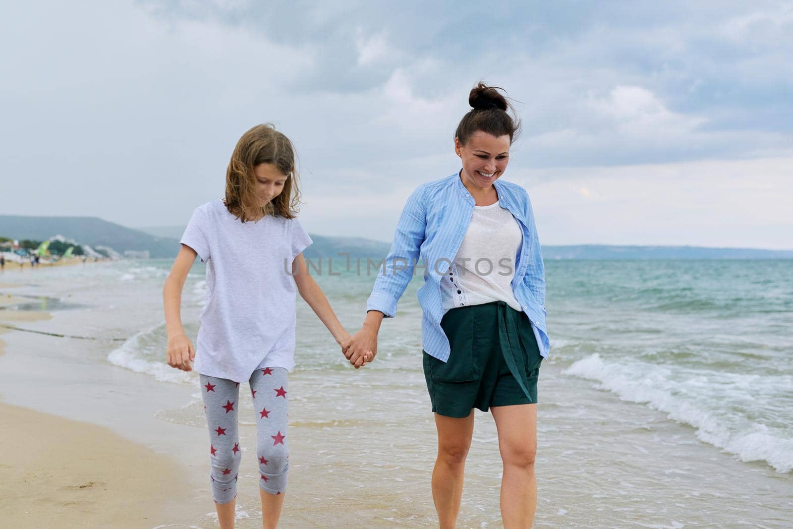 Happy mom and daughter walking on the sea beach holding hands. Family middle-aged mother and preteen child together, vacation, sea weekend, travel, love, happiness and joy