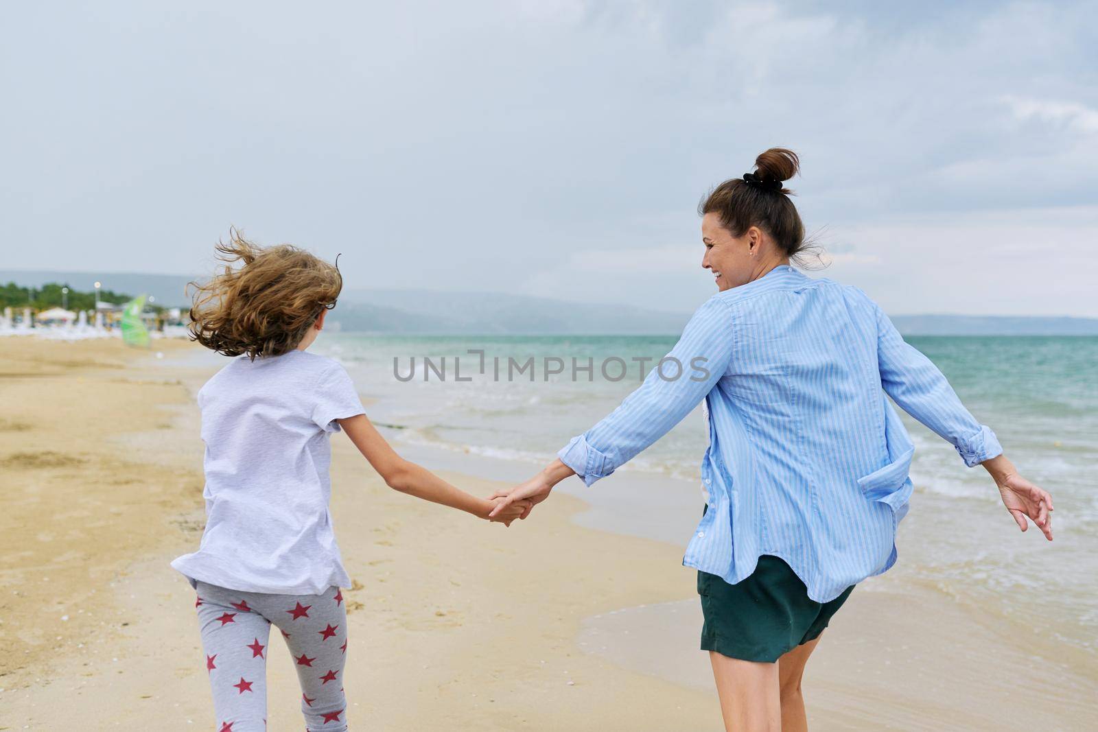 Happy mom and daughter running on sea beach holding hands, back view. Family middle-aged mother and preteen child together, vacation, sea weekend, travel, love, leisure, happiness and joy