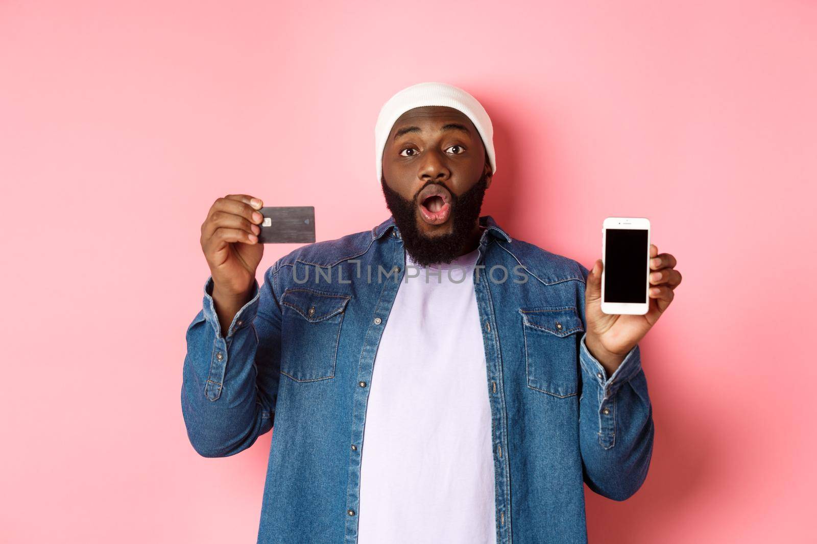 Online shopping. Excited Black man showing credit card and mobile phone screen, standing over pink background amazed.