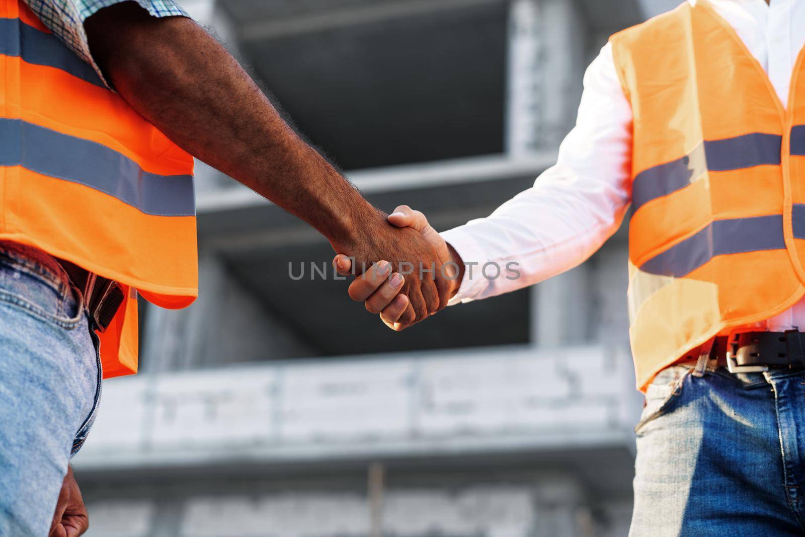 Two men engineers in workwear shaking hands against construction site, close up