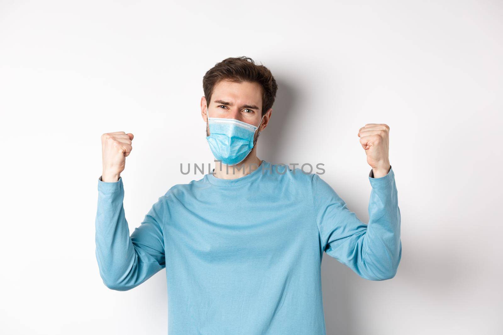 Covid-19, pandemic and social distancing concept. Happy young man in medical mask winning, screaming yes with satisfaction and raising hands up, celebrating victory, white background.