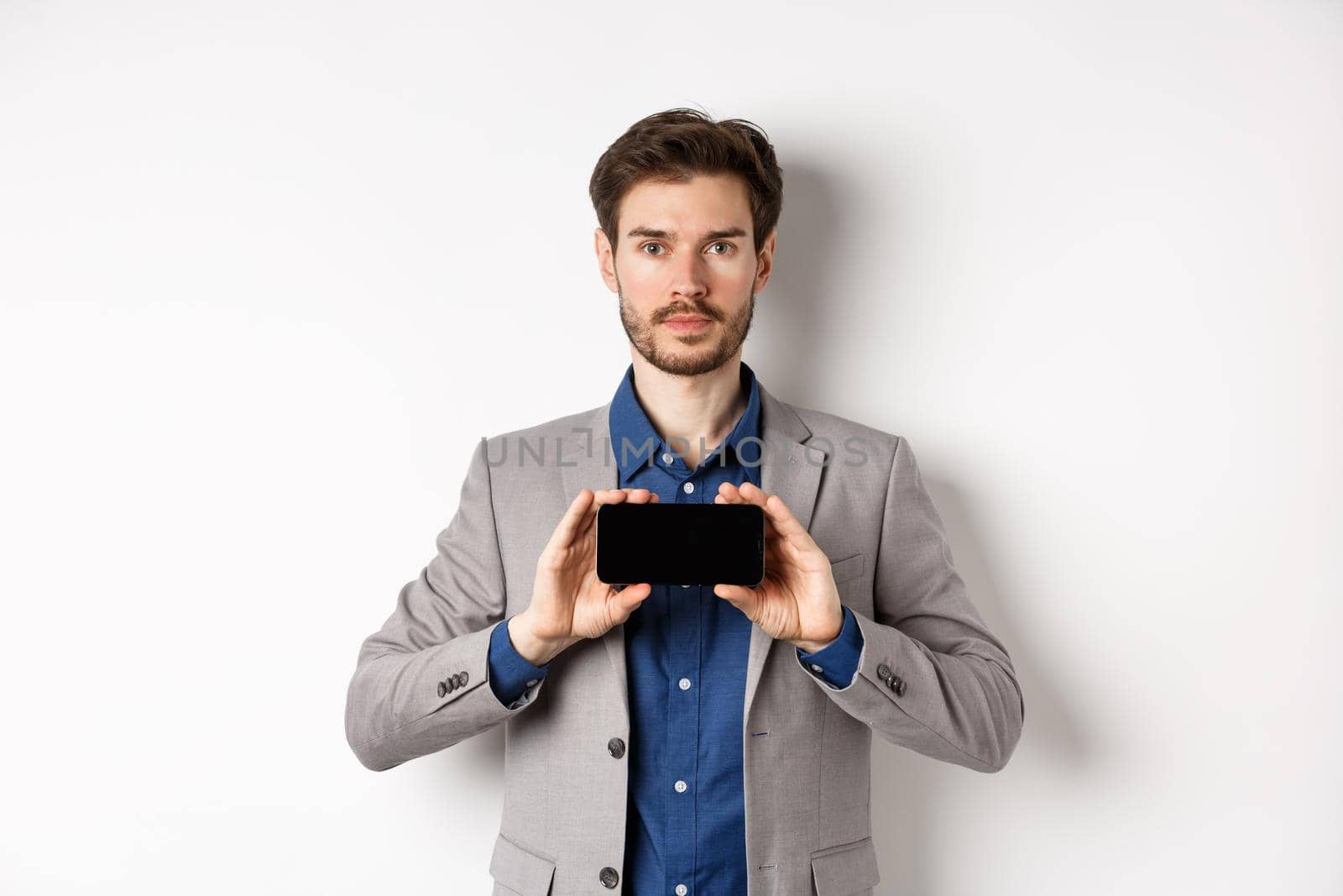 E-commerce and online shopping concept. Serious young man in suit showing empty smartphone screen horizontal, standing on white background.