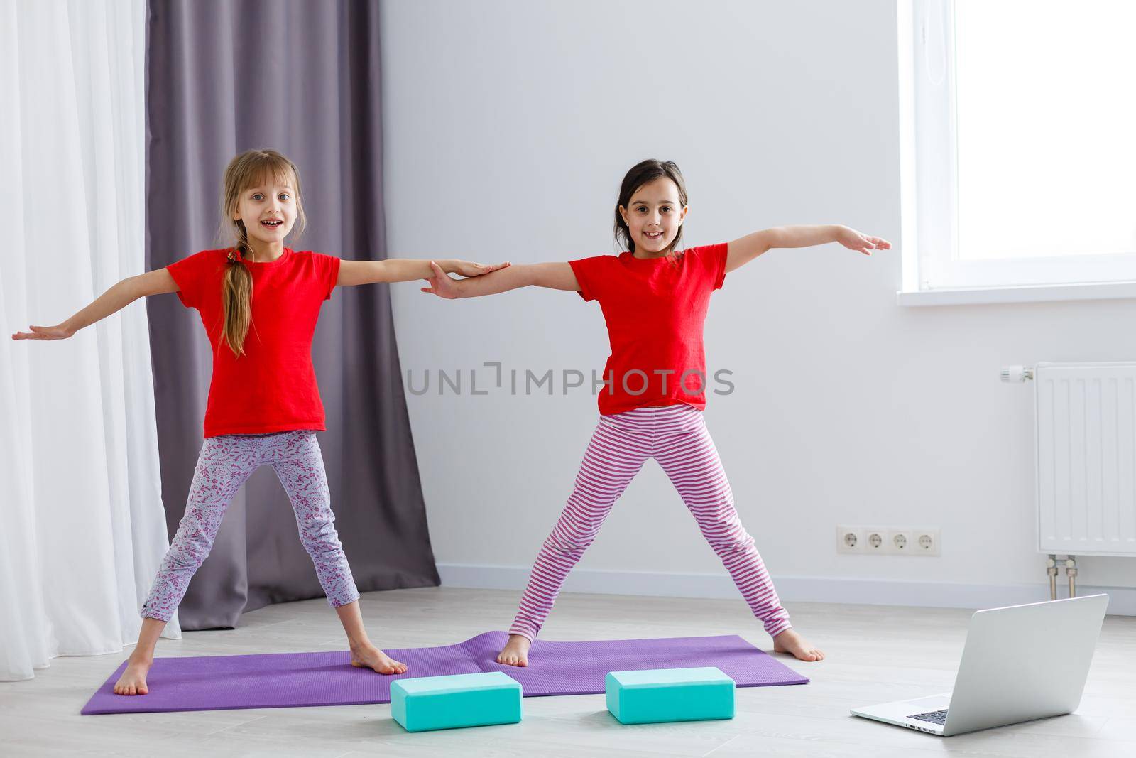 two little girls practicing yoga, stretching, fitness by video on notebook. Distant online education training, aerobic at home. Healthy lifestyle, coronavirus, stay home. Kids sport home quarantine. by Andelov13
