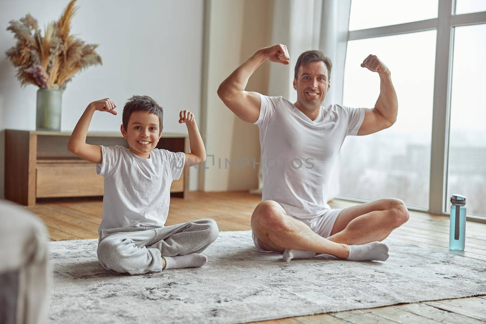 Full length portrait of happy muscular male sitting on mat with son and showing biceps together