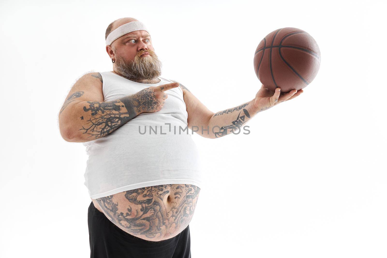 bearded fat caucasian tattoed man with basketball ball in white background. He is funny and happy