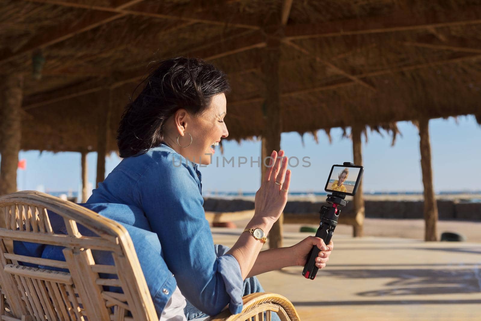 Middle aged woman looking at smartphone webcam talking recording video on sandy beach. Female blogger vlogger using video call on smartphone for blog, online stream, natural sea resort landscape