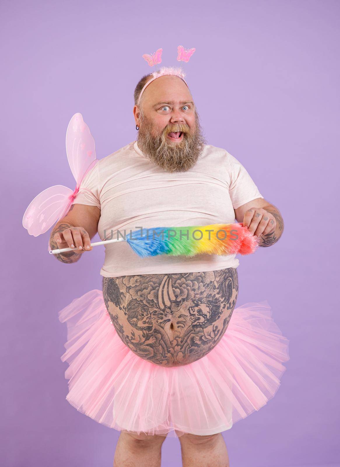 Bearded plump man in fairy costume holds colorful pp duster on purple background by Yaroslav_astakhov