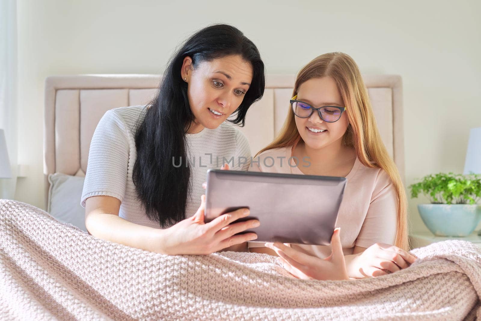 Happy mom and preteen daughter hugging together looking at screen of digital tablet, sitting at home in bed under blanket. Family, children, relationships, home, lifestyle, people concept