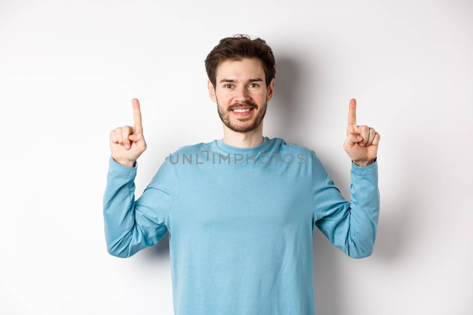 Image of young modern guy with beard showing advertisement on copy space, pointing fingers up and smiling, check it out gesture, white background.