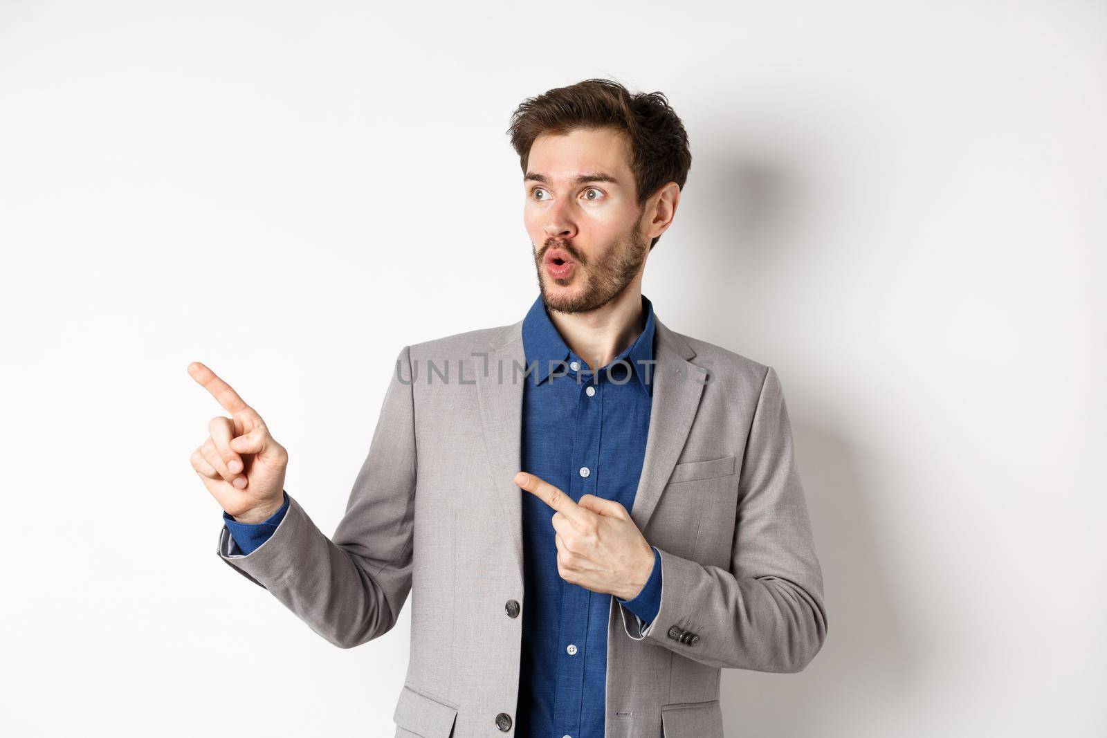 Wow look there. Excited male ceo manage in suit pointing fingers left, looking at logo impressed, showing advertisement, standing on white background by Benzoix