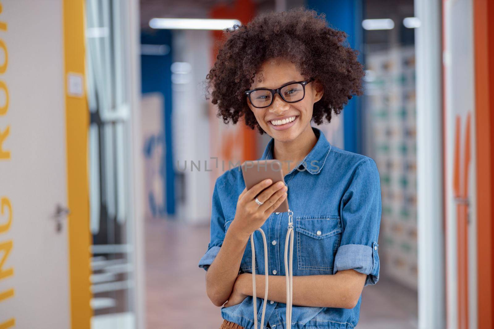 Portrait of happy Afro American lady in glasses standing in office corridor while using smartphone