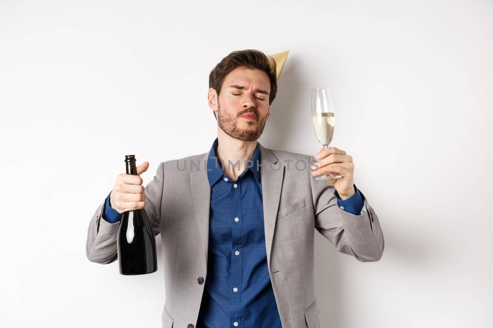 Celebration and holidays concept. Drunk guy having fun at birthday party, dancing with glass of champagne and bottle, wearing suit with cone hat, white background by Benzoix