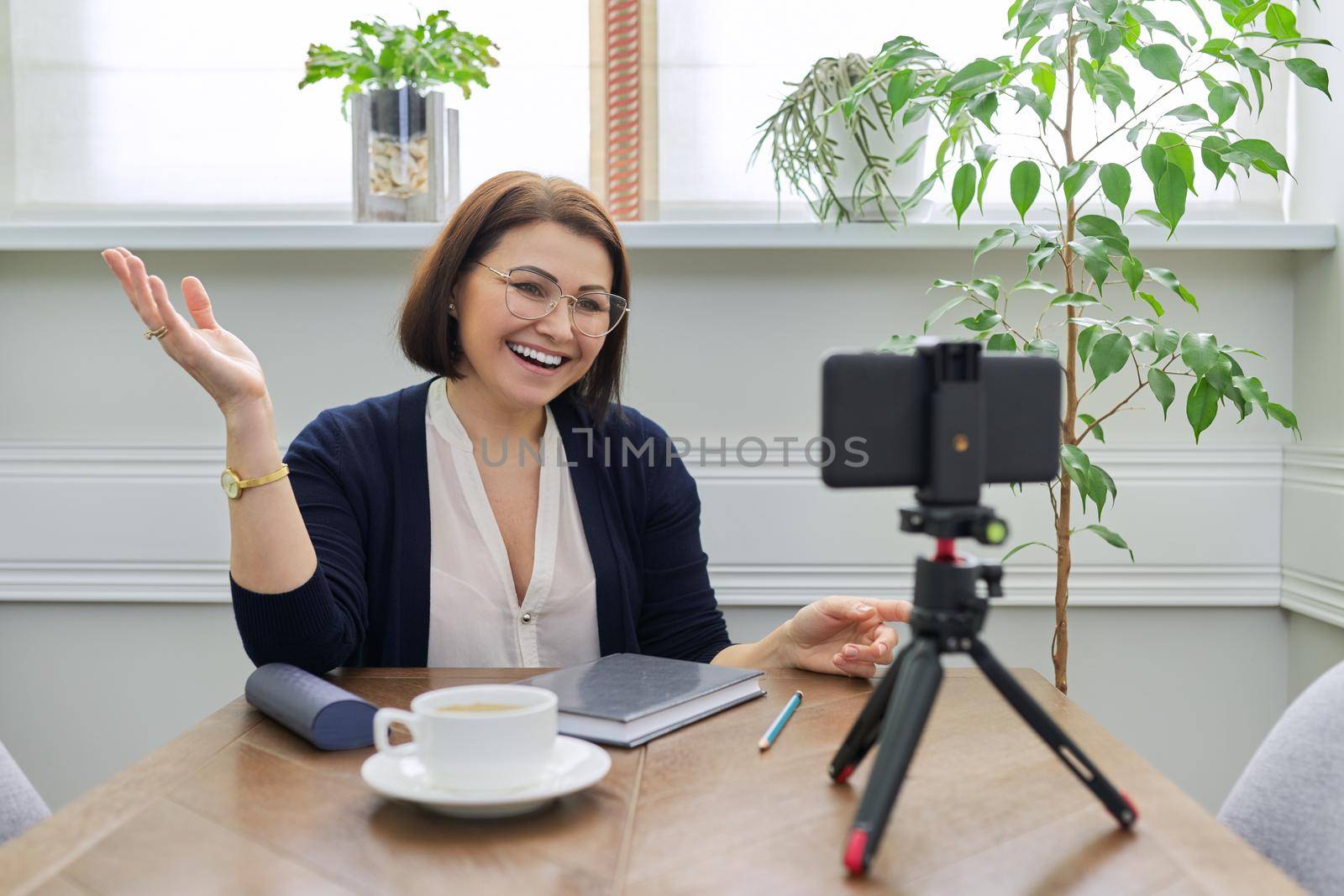 Woman teacher, business, psychologist, blogger, lawyer talking online using smartphone. Female looking at phone webcam on tripod, background table with cup of coffee notepad, window