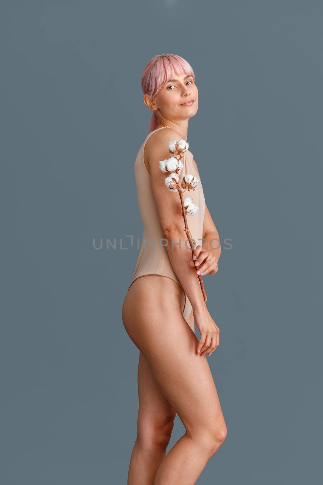 Beautiful woman with pink hair in beige bodysuit looking at camera, holding sprig with fluffy cotton flowers, standing isolated over gray studio background. Naturalness, beauty concept