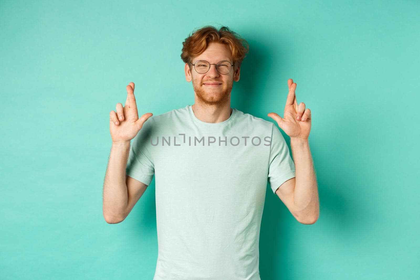 Young cheerful guy with red hair and beard, wearing glasses, smiling and cross fingers for good luck, making wish and looking optimistic, standing over mint background by Benzoix
