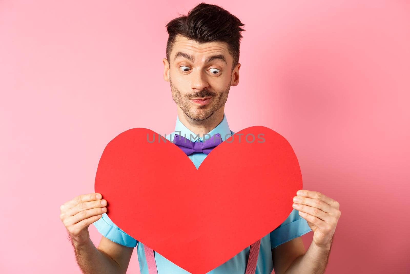 Valentines day concept. Cute boyfriend looking at big red heart, waiting for lover on romantic date, pink background.
