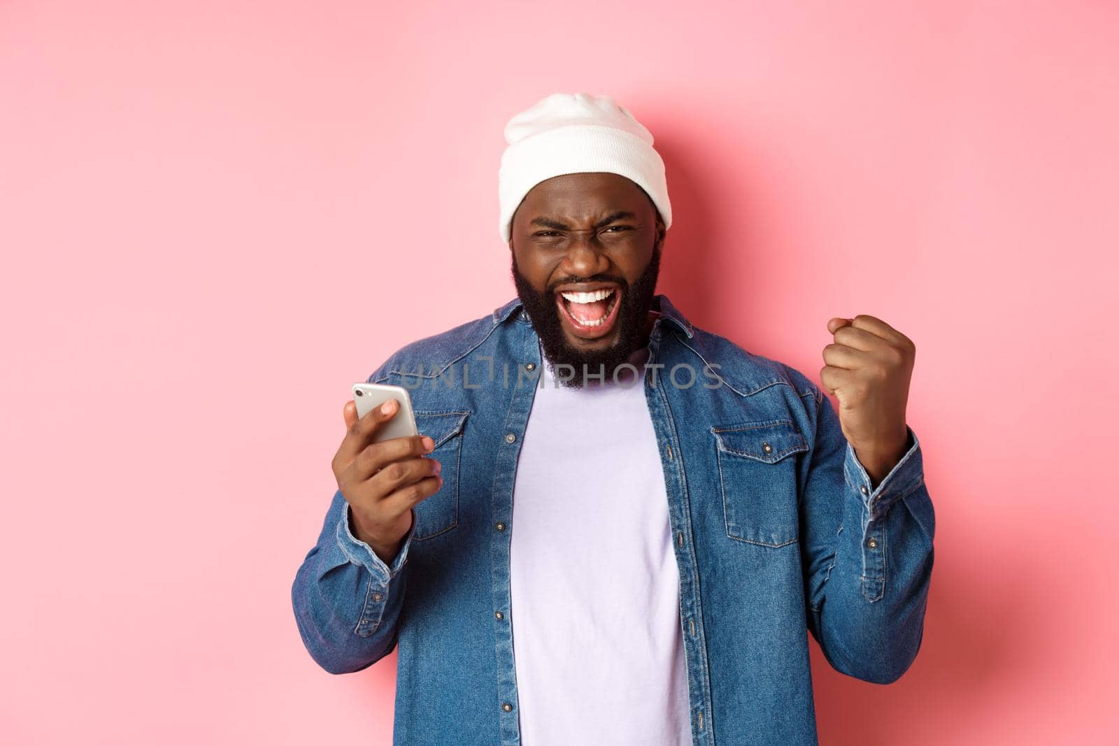 Technology and online shopping concept. Happy Black man rejoicing, winning in app, holding smartphone and scream yes, standing over pink background.