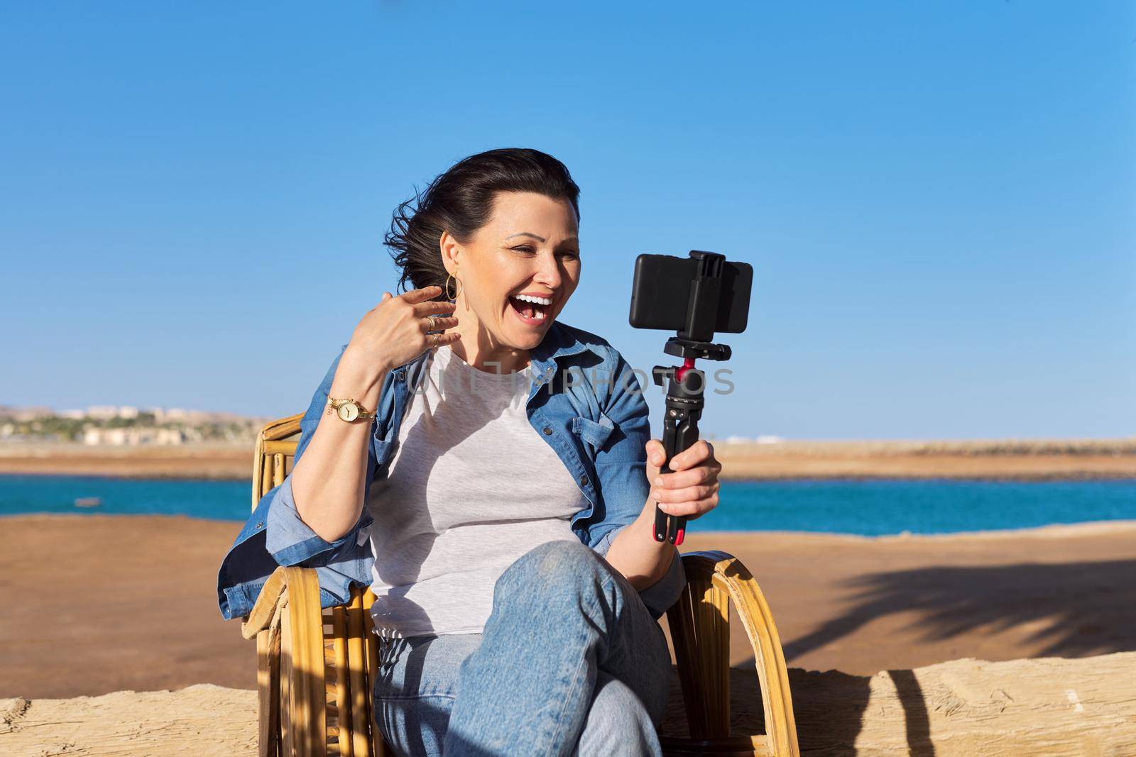 Middle aged beautiful woman recording video on smartphone, sunset sea beach background. Female blogger, vlogger, talking to followers using video call, recording stream