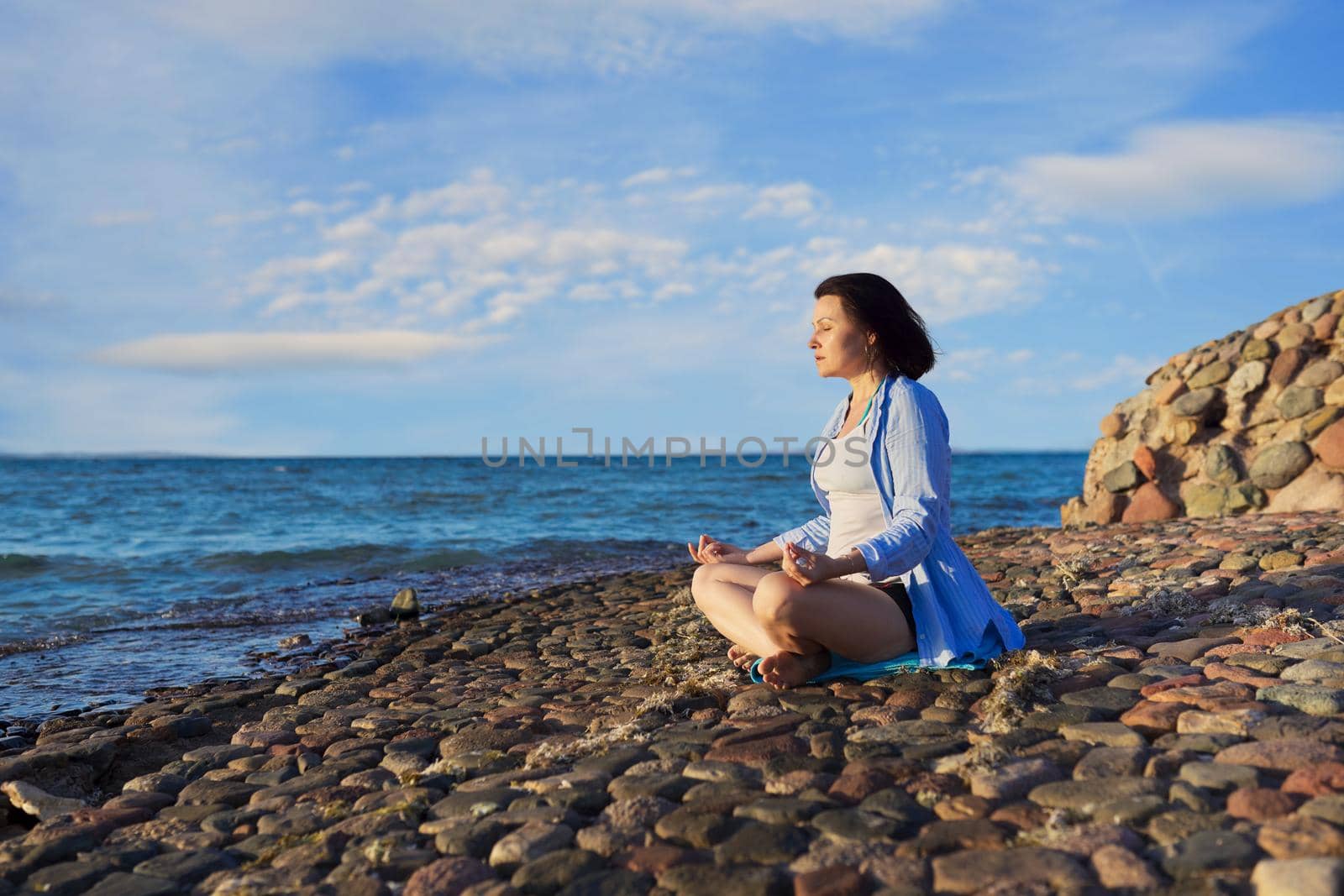 Beautiful middle aged woman sitting in lotus position meditating on seashore at sunset by VH-studio