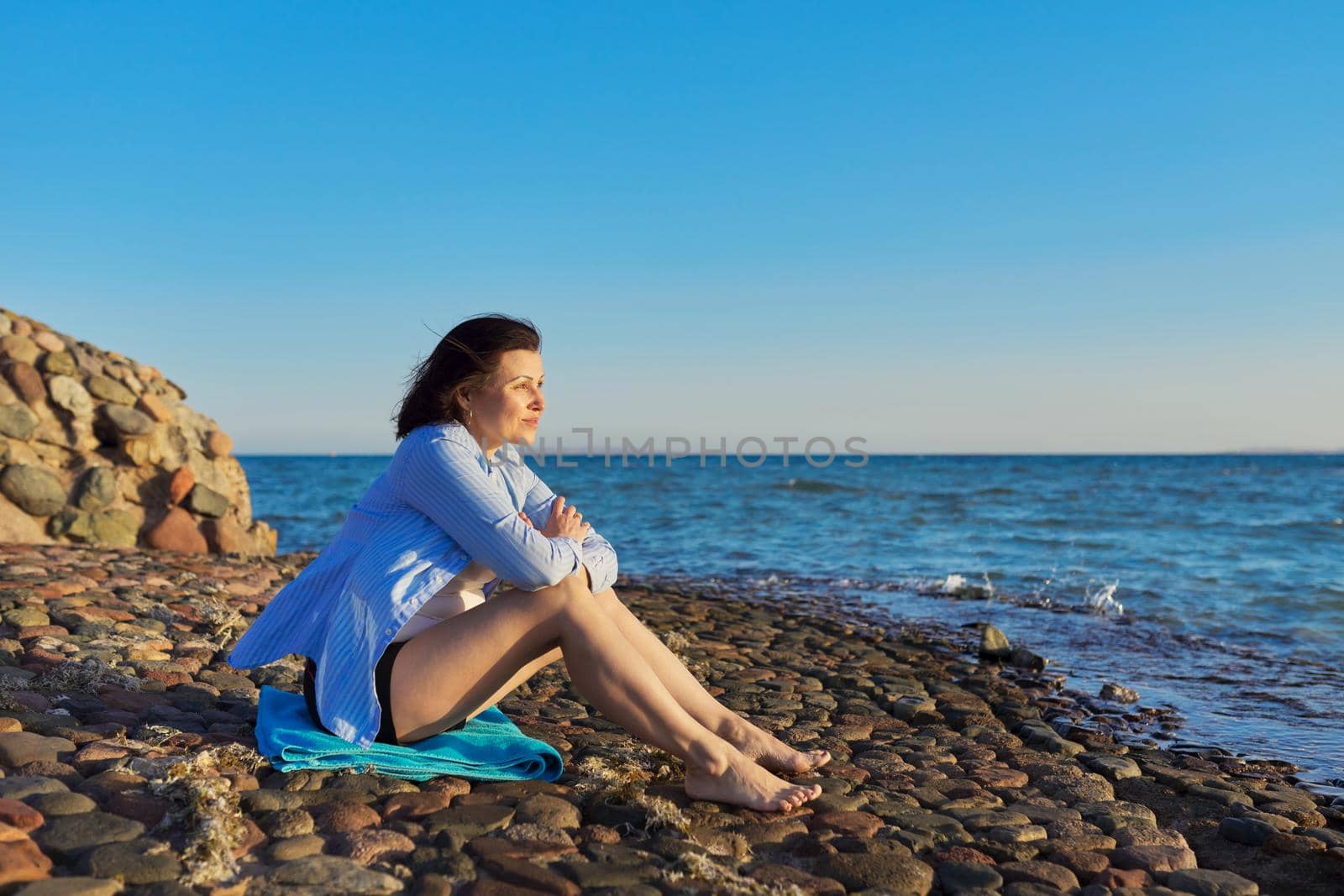 Mature woman sitting on the seashore enjoying the seascape, nature, sunset. Meditating, relaxing, breathing sea air, copy space