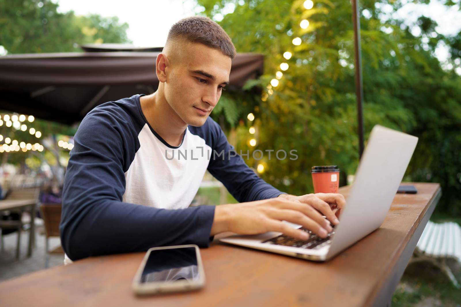 Young man sitting at table and typing on laptop keyboard while working in outdoor cafe, close up