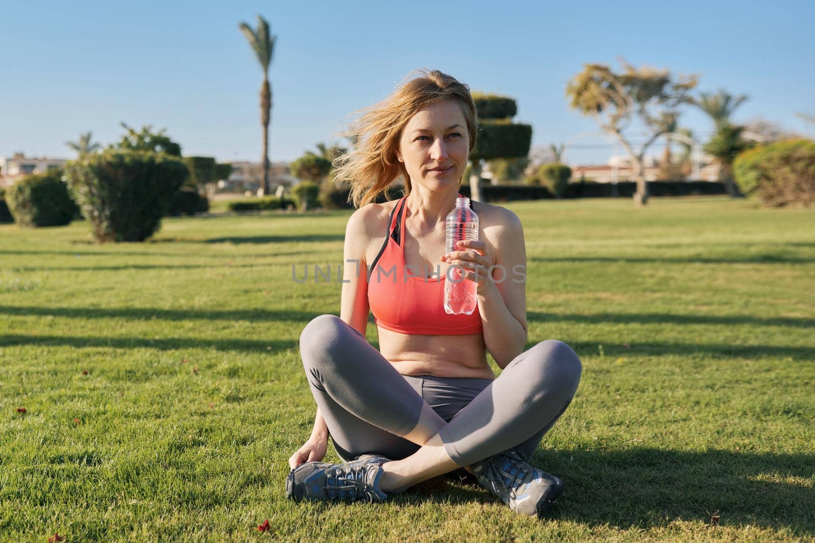 Mature woman doing morning exercises on green grass in park, with a bottle of water, sports, healthy active lifestyle of middle-aged people