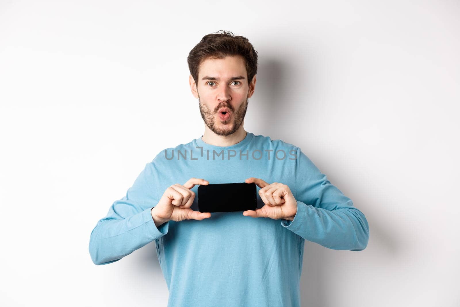 E-commerce and shopping concept. Image of impressed man saying wow and showing blank mobile phone screen in horizontal, standing over white background.