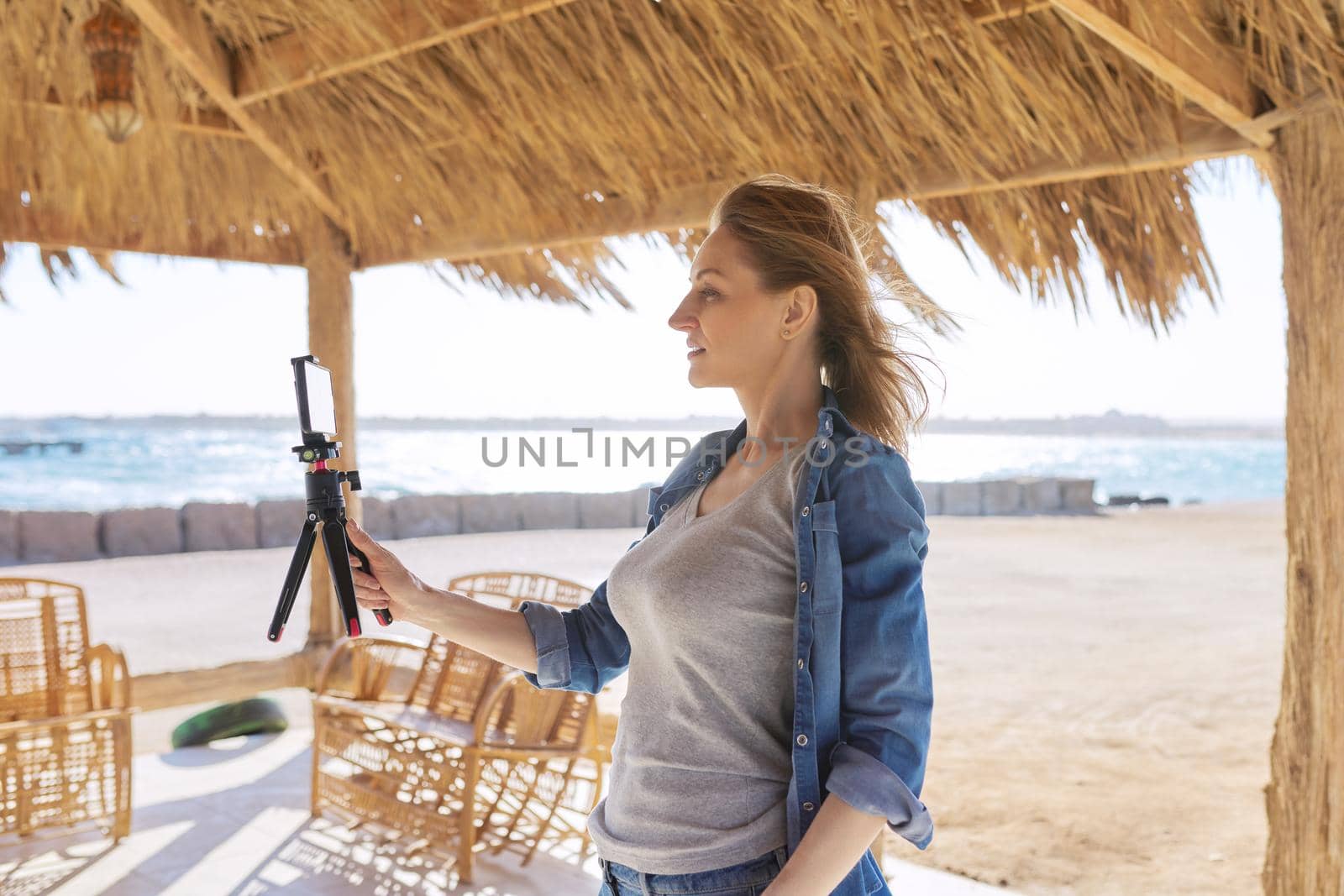 Middle aged woman looking at smartphone webcam talking recording video on sandy beach. Female blogger vlogger using video call on smartphone for blog, online stream, natural sea resort landscape