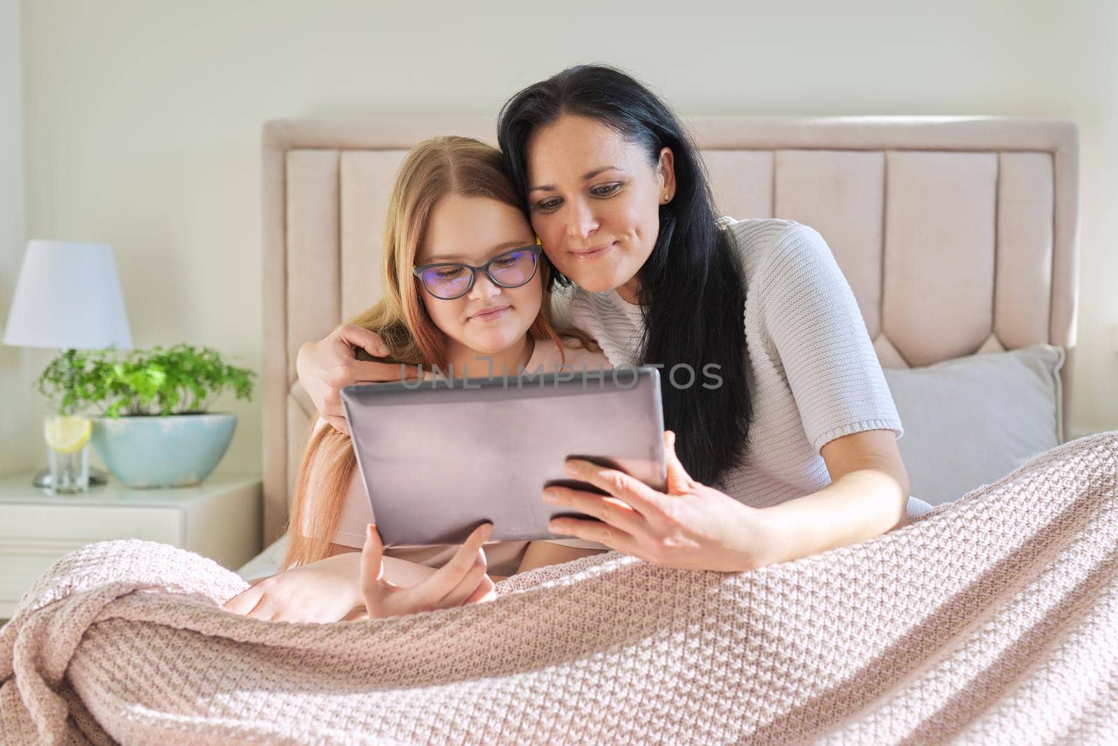 Happy mom and preteen daughter hugging together looking at screen of digital tablet, sitting at home in bed under blanket. Family, children, relationships, home, lifestyle, people concept