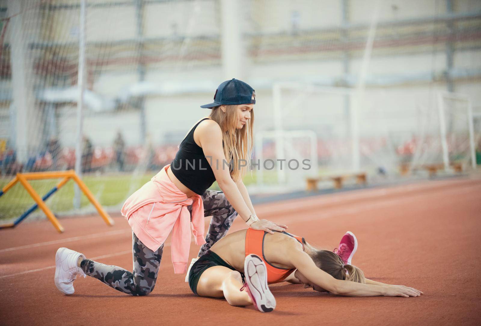 Young beautiful woman with blonde hair helps another woman to stretch her legs in the sports hall after training by Studia72
