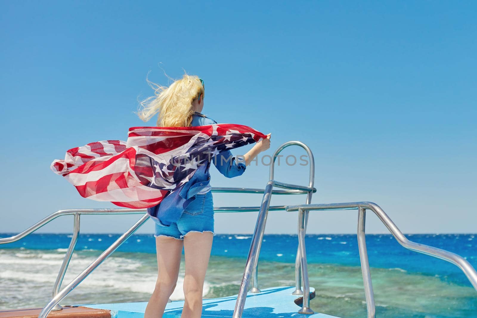 Young slender blonde woman in denim clothes with the flag of America on her shoulders, back view, in the sea on a yacht