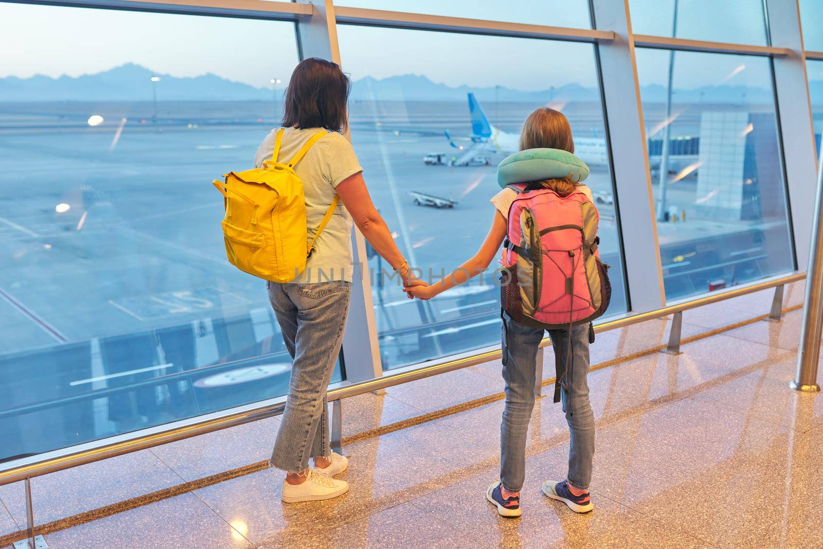 Airport passengers, standing with their backs family mother and daughter child looking at planes in panoramic window, awaiting boarding, copy space