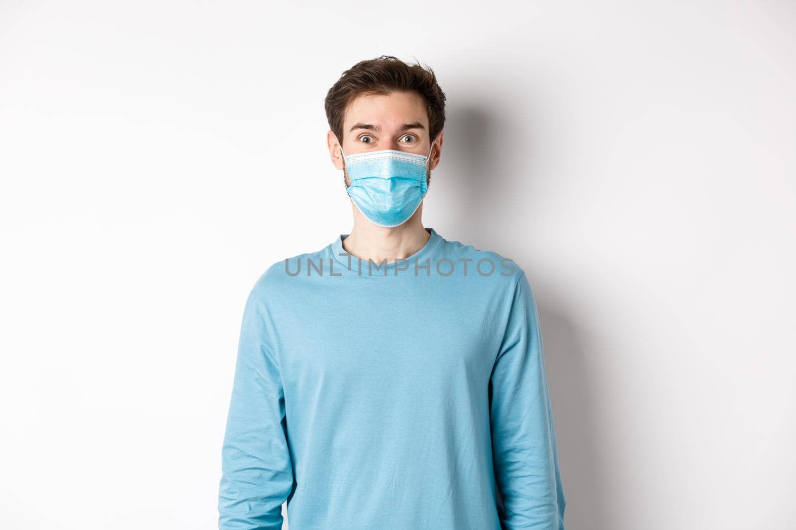 Covid-19, pandemic and social distancing concept. Cheerful man in medical mask raising eyebrows, looking surprised and excited at camera, standing over white background.