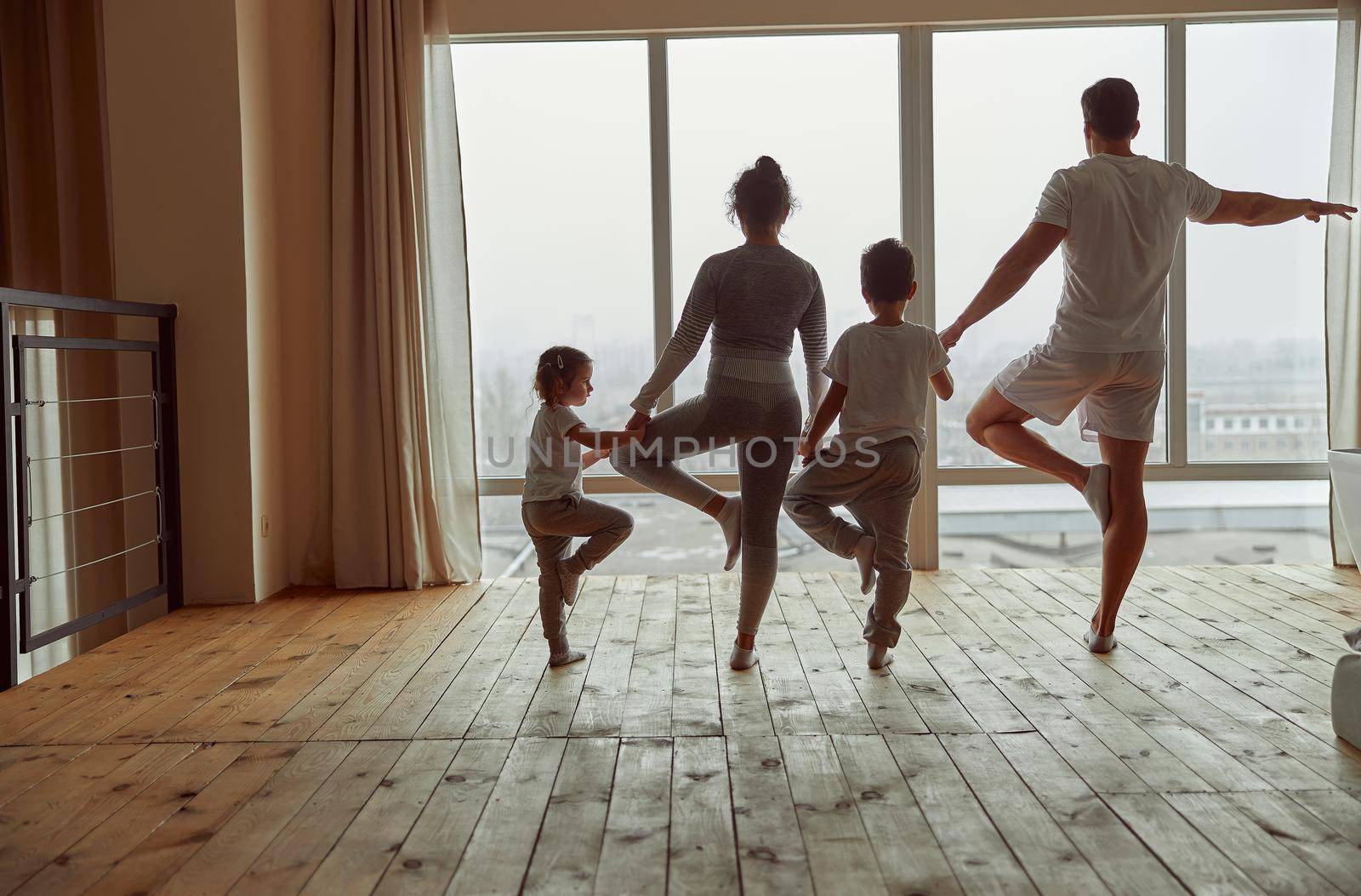 Sporty parents with son and daughter are standing on one foot in asna with faces to window