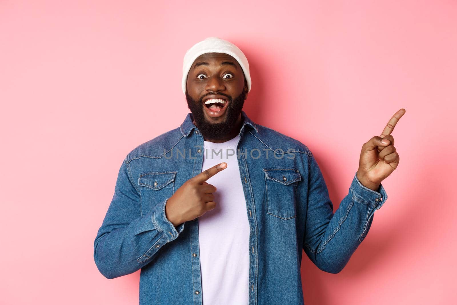 Excited and amazed Black man showing awesome offer, pointing fingers right at copy space, standing in hipster beanie and denim shirt on pink background.