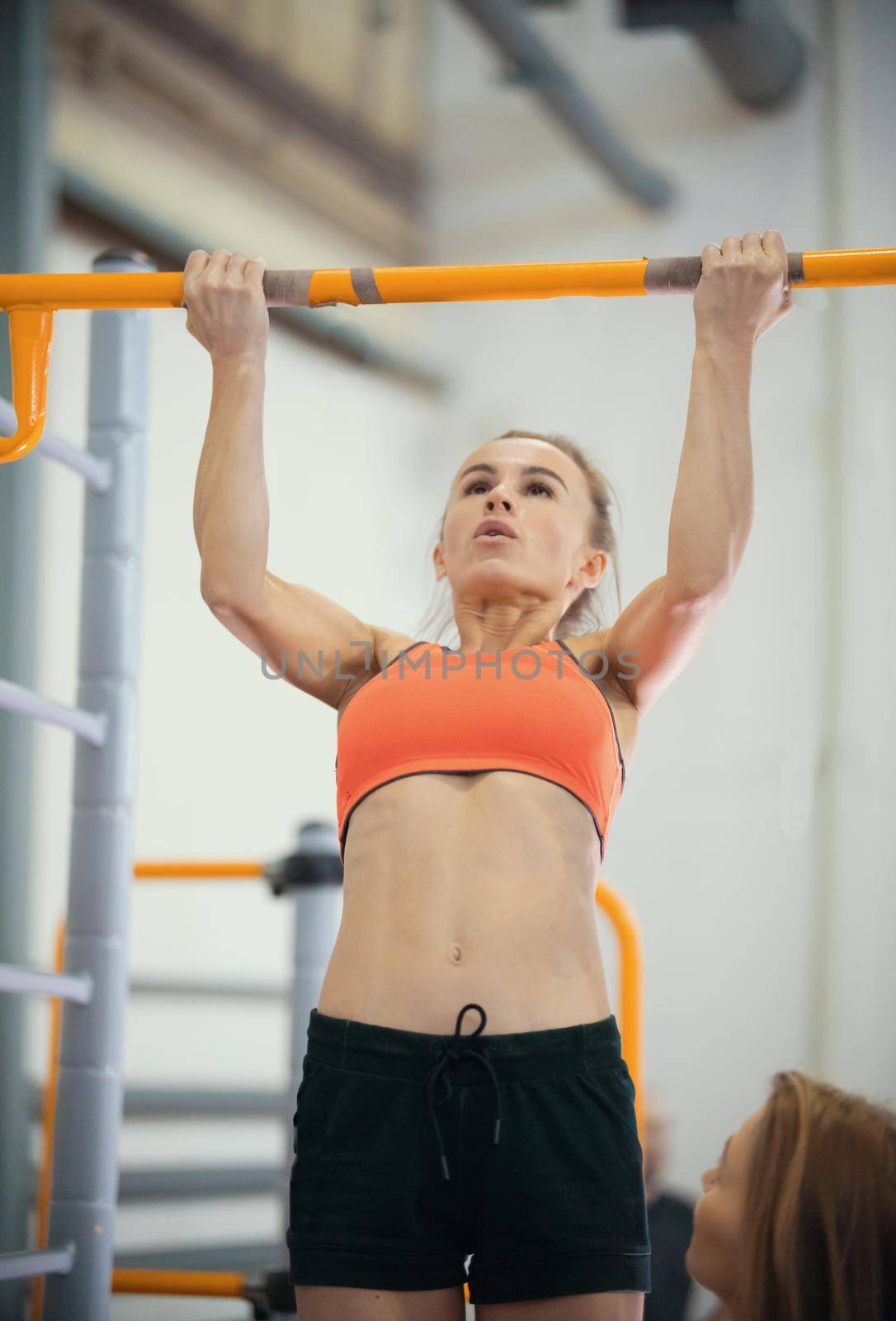 Young woman with blonde hair and strong arms pulls up on the crossbar in the sports hall