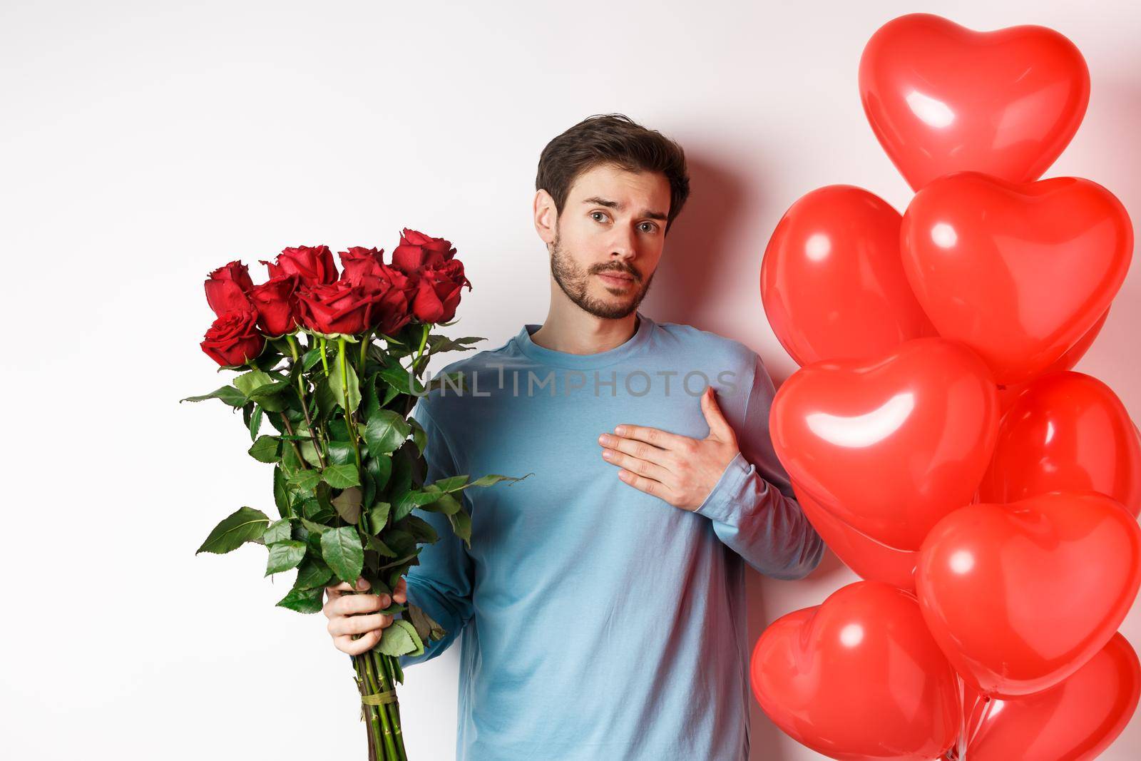 Romantic guy express his love on Valentines day with gifts, bring bouquet of red roses and balloons, holding hand on heart, standing over white background.