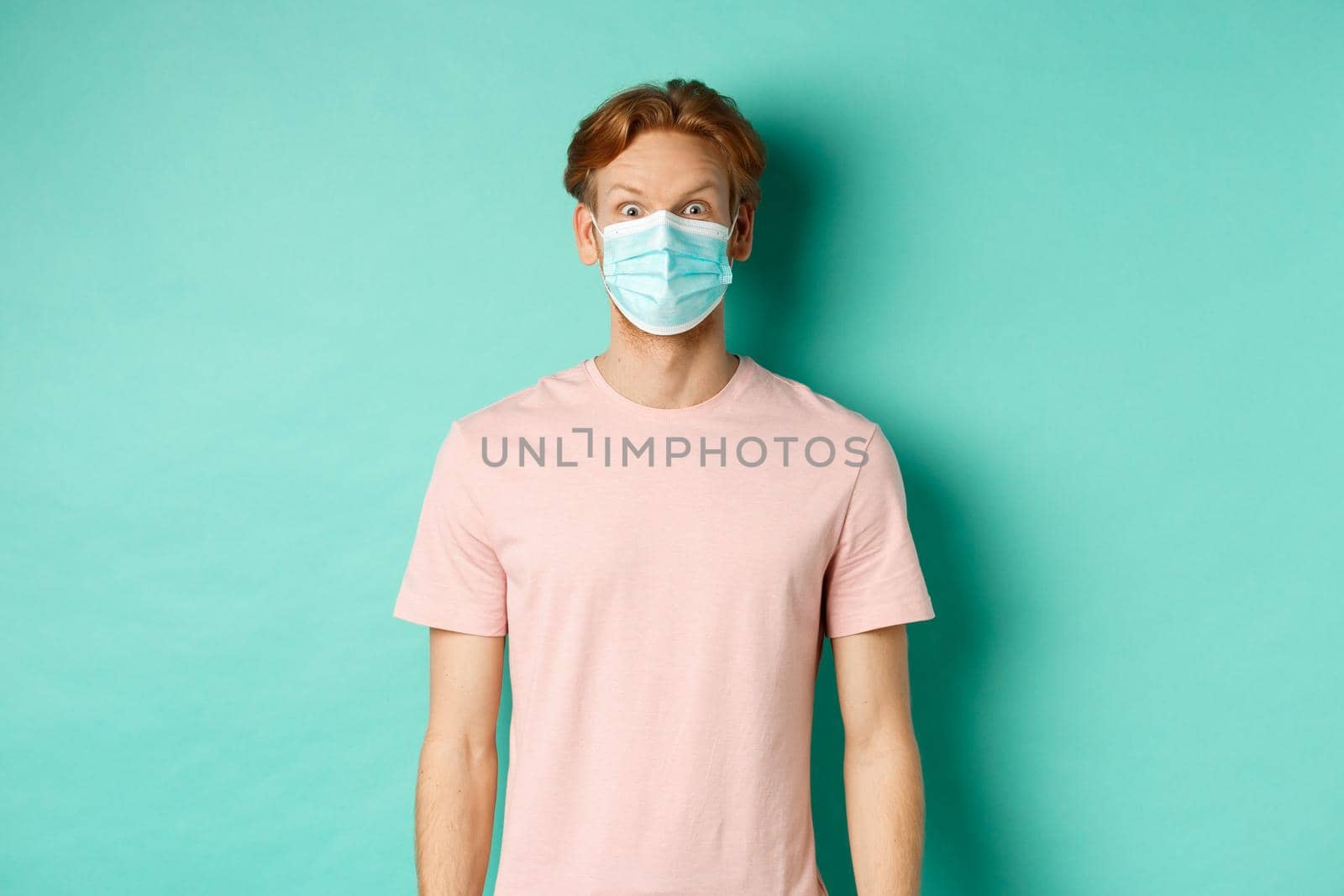 Covid-19, social distancing and quarantine concept. Young man raising eyebrows and looking surprised, wearing face mask during pandemic, standing over mint background.