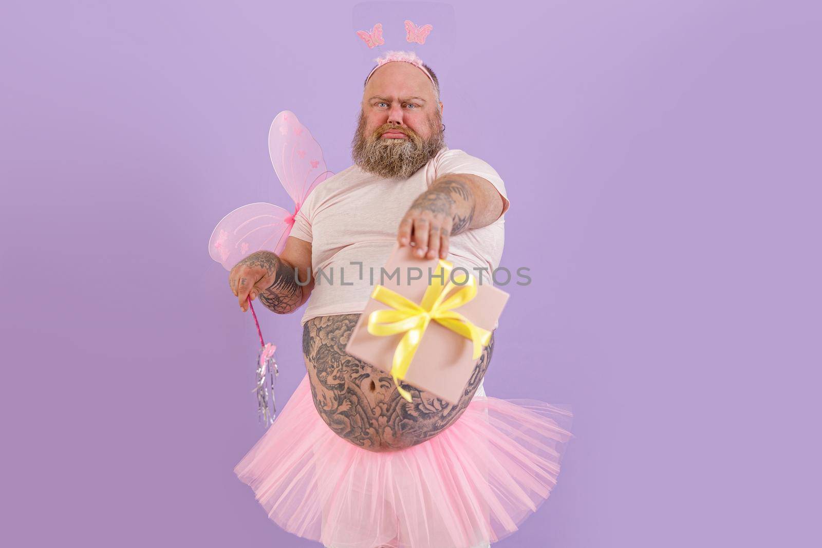 Emotional brutal obese man wearing fairy costume holds magic stick and gift box grimacing on purple background in studio