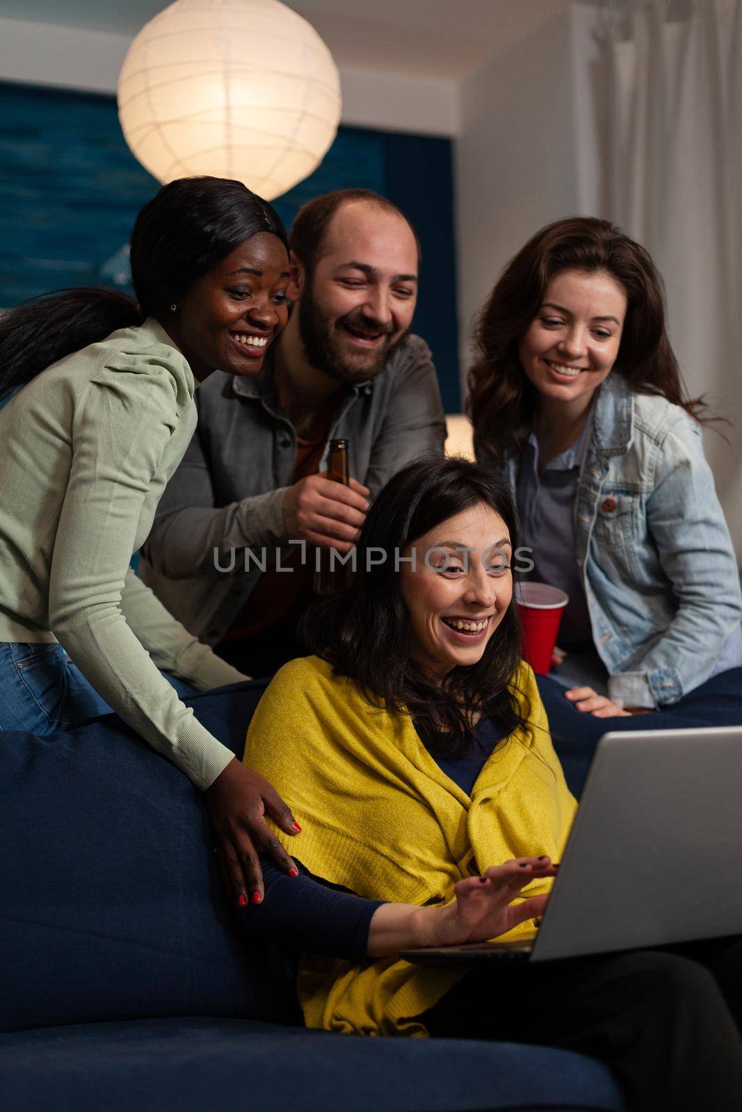 Multi-ethnic friends relaxing on couch laughting while watching fuuny comedy film during movie night at home enjoying spending time together. Group of people hanging out in evening. Friendship concept