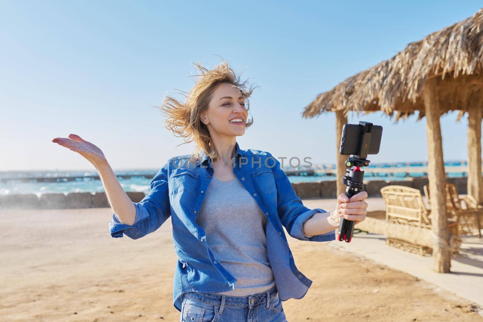 Middle aged woman looking at smartphone webcam talking recording video on sandy beach. Female blogger vlogger using video call on smartphone for blog, online stream, natural sea resort landscape