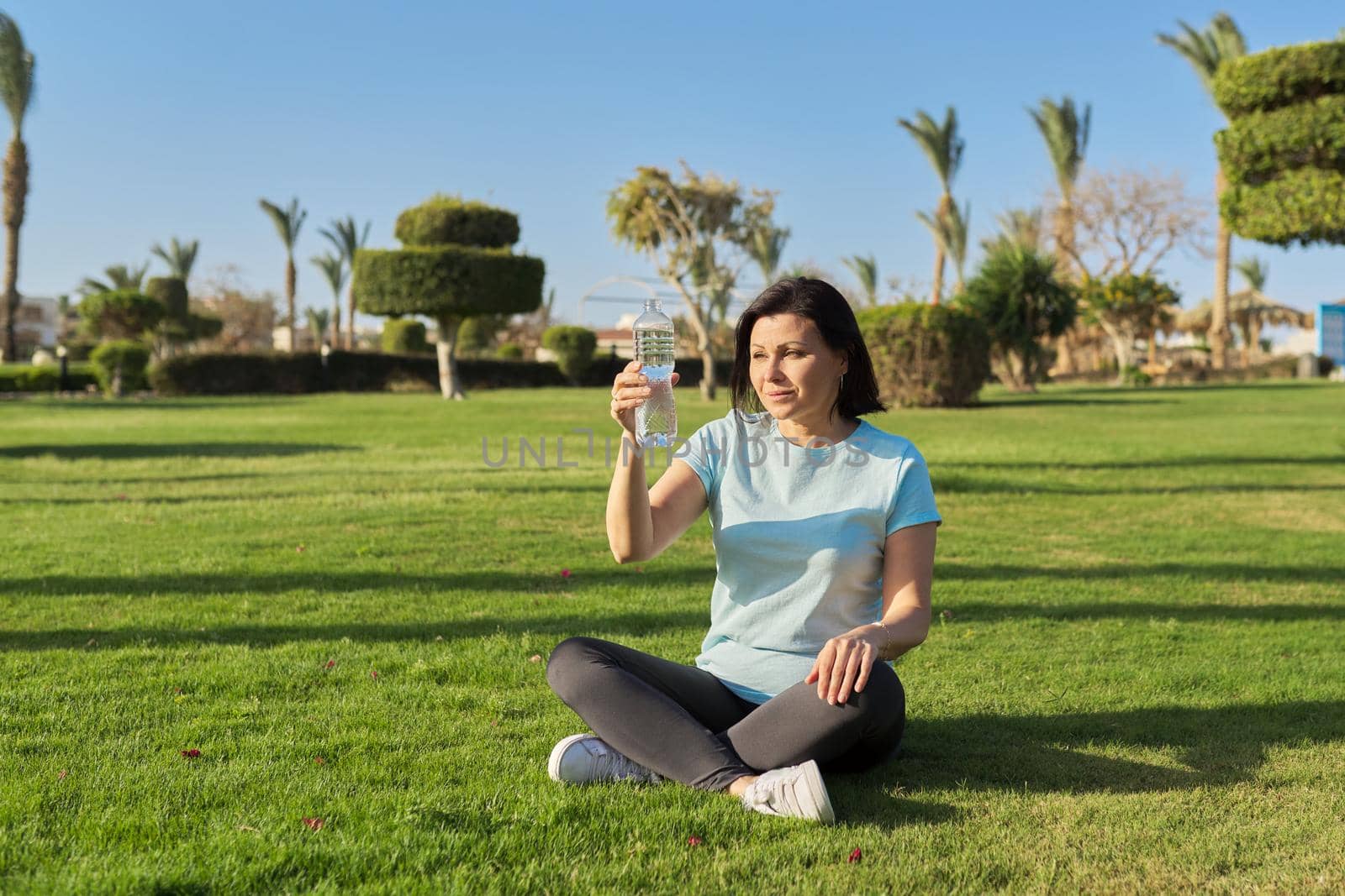 Mature woman doing morning exercises on green grass in park, with bottle of water by VH-studio