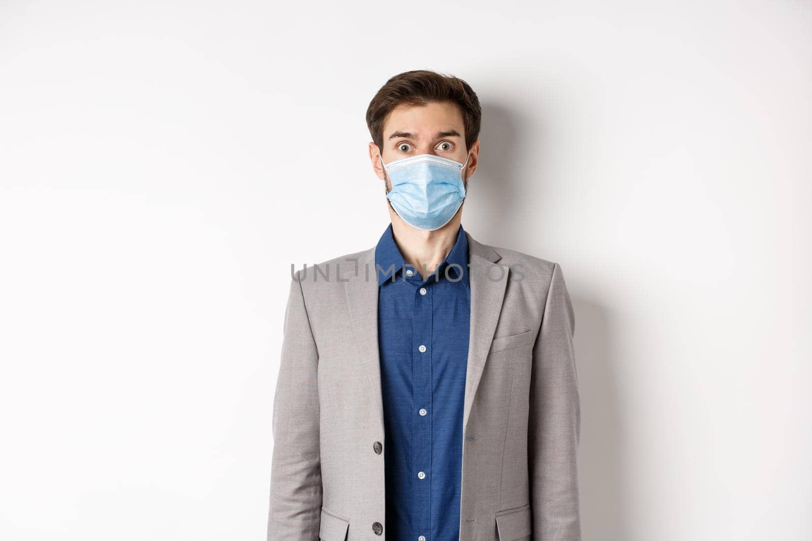 Health, pandemic and business concept. Surprised guy in suit and medical mask raising eyebrows, stare shocked at camera, white background by Benzoix