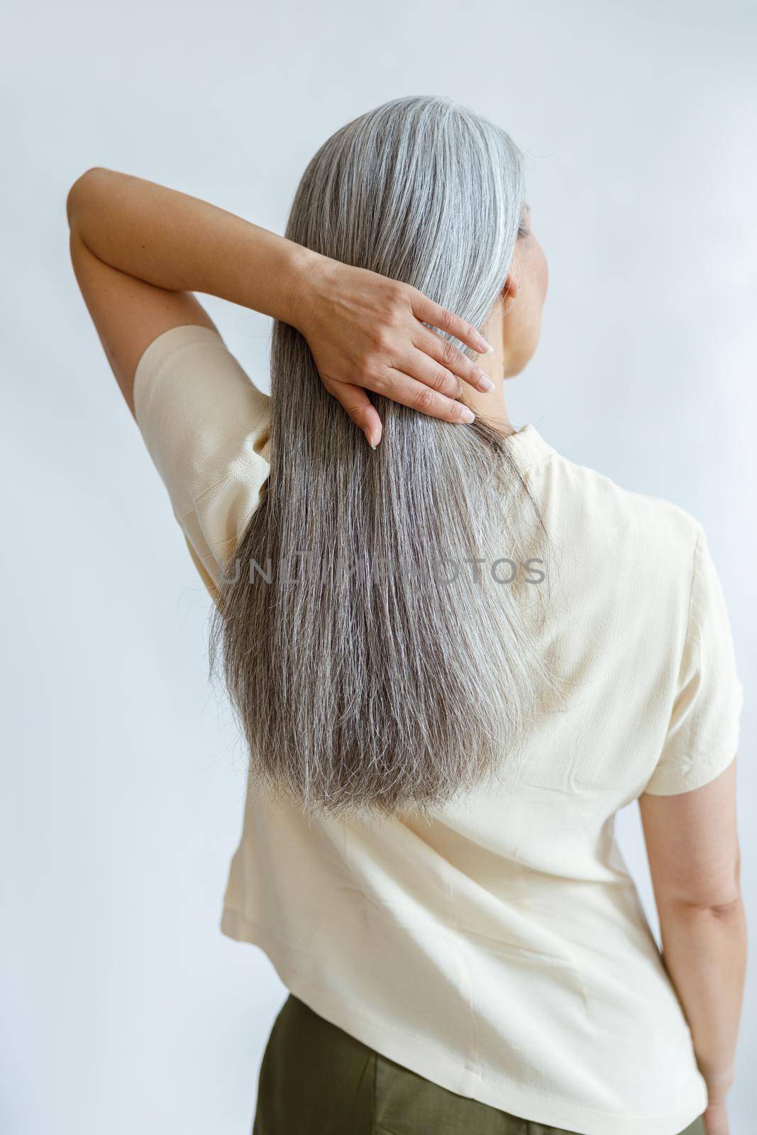 Middle aged woman adjusts long loose hoary hair on light grey background by Yaroslav_astakhov